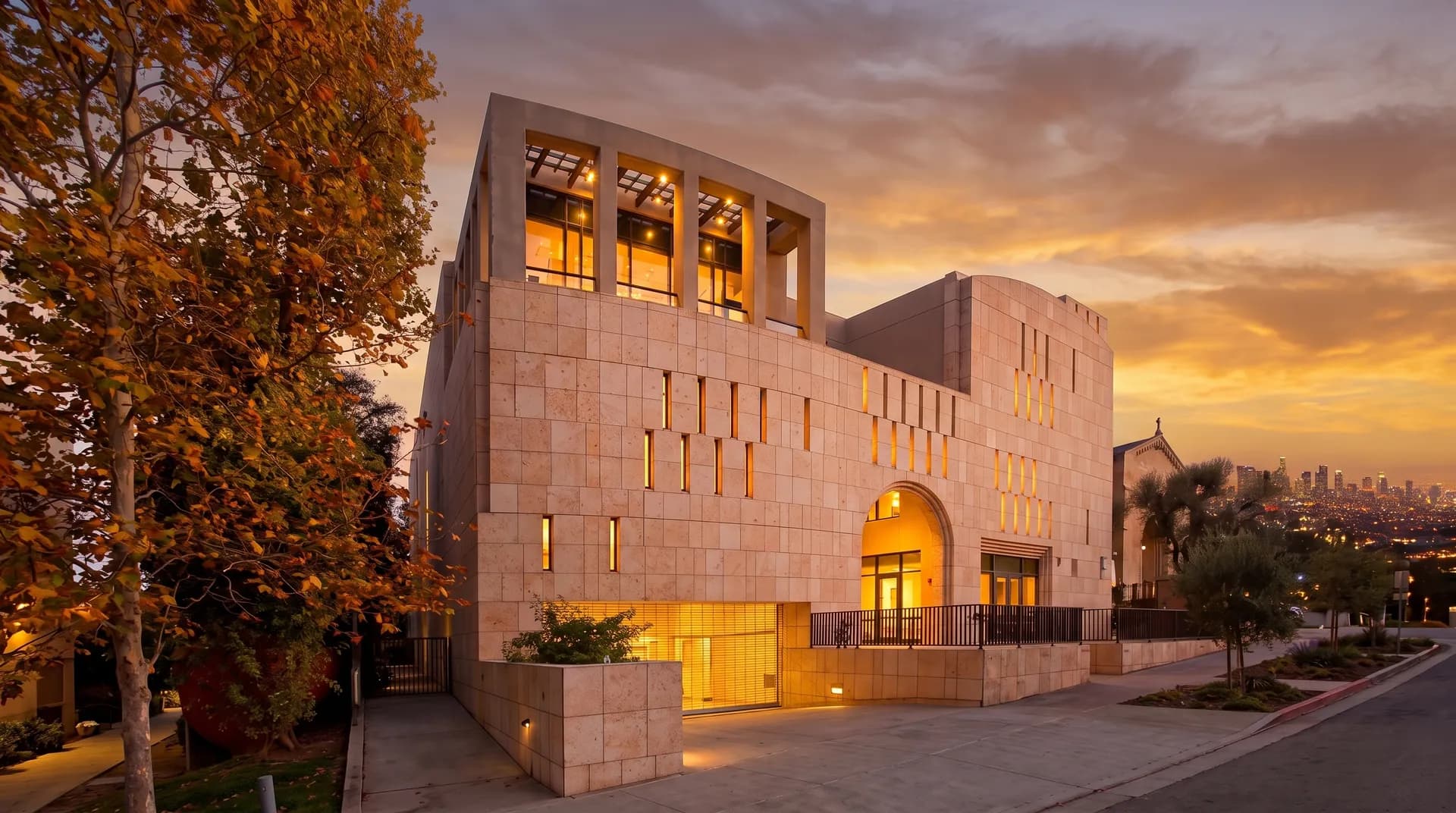 Modern travertine stone facade, vertical window slits, upper pergola terrace, and arched entryway of Hillel at UCLA at 574 Hilgard Avenue in rich golden hour sunset light with glowing illuminated windows.