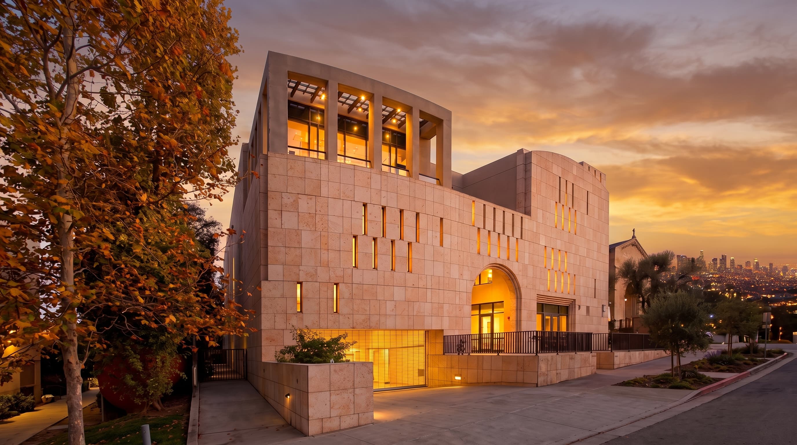 Modern travertine stone facade, vertical window slits, upper pergola terrace, and arched entryway of Hillel at UCLA at 574 Hilgard Avenue in rich golden hour sunset light with glowing illuminated windows.